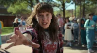 Millie Bobby Brown as Eleven in season four of Stranger Things. She has longer hair and bangs, wearing flannel and a t-shirt and stretching her right arm out to use her powers.