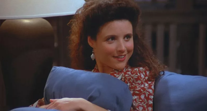 Julia Louis-Dreyfus smiling and sitting on the couch as Elaine Benes on Seinfeld