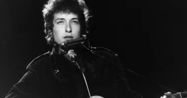 A black and white picture of Bob Dylan playing guitar and standing in front of a microphone.
