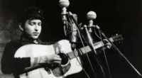 A black and white picture of Bob Dylan playing guitar in front of three microphones.
