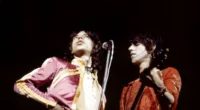 Mick Jagger stands behind a microphone with his hand on his hip. Keith Richards stands next to him, playing guitar.