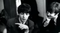 Paul McCartney (left) pointing to his right while John Lennon watches before a December 1963 concert in London.