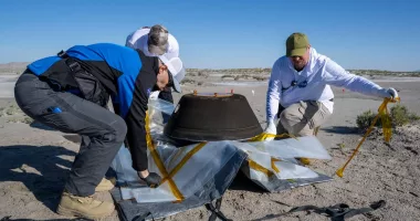 Three members of NASA circled around the OSIRIS-REx capsule that landed in the desert, preparing it for transport.