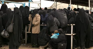 A number of women dressed in black hijabs waiting for supplies outside a Syrian camp. One of the crouched women holds a baby in her arms.
