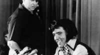 A black and white picture of Colonel Tom Parker wearing a cowboy hat and watching Elvis, who sits in front of multiple microphones.