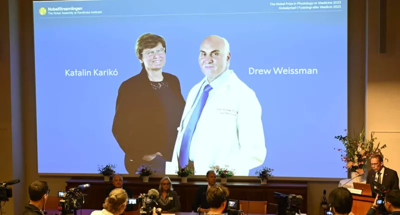 A man talks at a podium next to a table of people, with a blue screen showing Katalin Kariko and Drew Weissman behind them.
