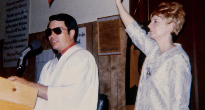 Jim Jones and his wife, Marceline preaching standing at a podium