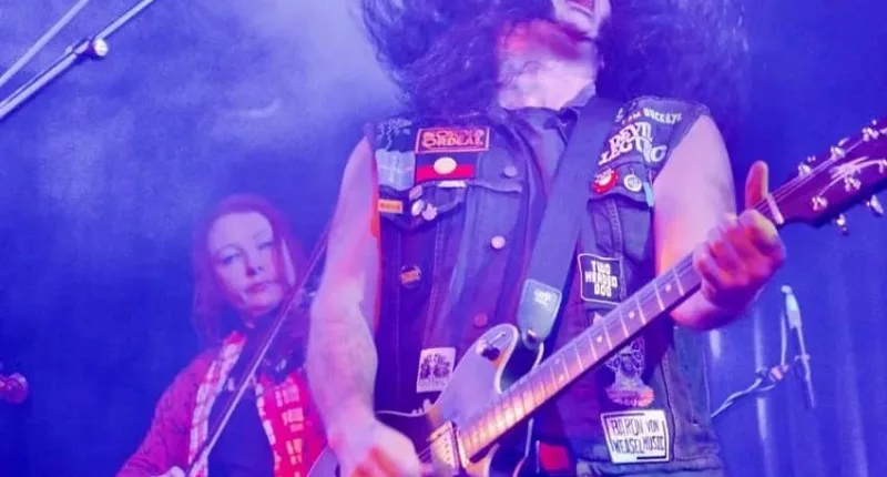 A man in a black denim vest covered in patches plays the electric guitar while flicking his long, dark hair. A woman plays a string instrument behind him