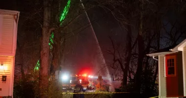Fire and police officials walk around the scene of a house explosion as an Arlington County Fire Department ladder truck sprays water down on the remains of the building on Monday, Dec. 4, 2023, in Arlington, Va. (AP Photo/Kevin Wolf)
