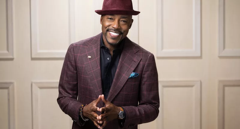A headshot of Will Packer wearing a purple had and suit