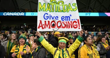 A crowd of people dressed in yellow and green. A man holds up a sign reading 'Go Matildas give em a-moosing'