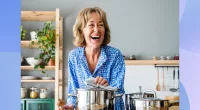 happy senior woman wearing blue button-down shirt laughing and cooking a healthy meal or soup in bright kitchen