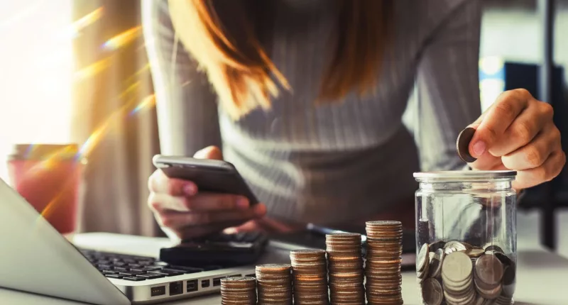 a lady holding a phone and a coin, coins on the table in a line up with some filled in a jar, laptop on the desk