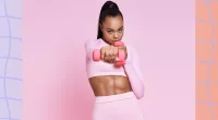 fit, focused woman in pink workout attire doing dumbbell exercise in front of pink backdrop