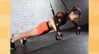 fit brunette woman wearing orange biker shorts and black sports bra doing TRX exercise at the gym
