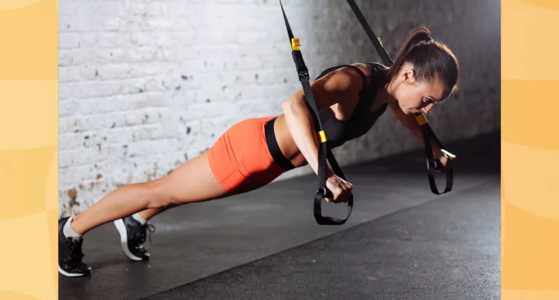 fit brunette woman wearing orange biker shorts and black sports bra doing TRX exercise at the gym