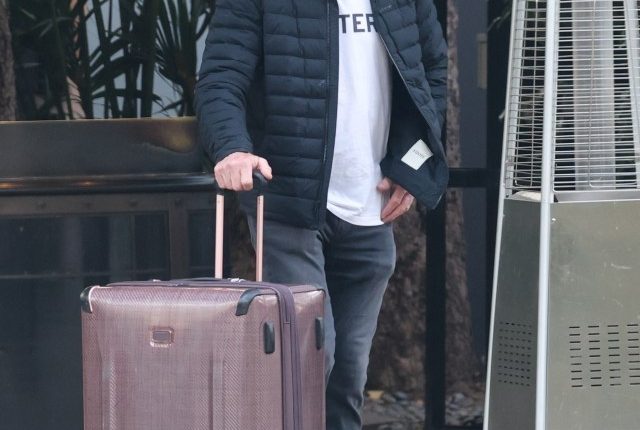Dennis Quaid pulling a suitcase outside a hotel.