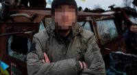 British frontline medic with face obscured, standing in front of damaged military vehicle.