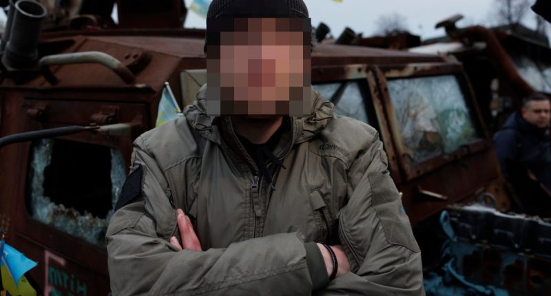 British frontline medic with face obscured, standing in front of damaged military vehicle.
