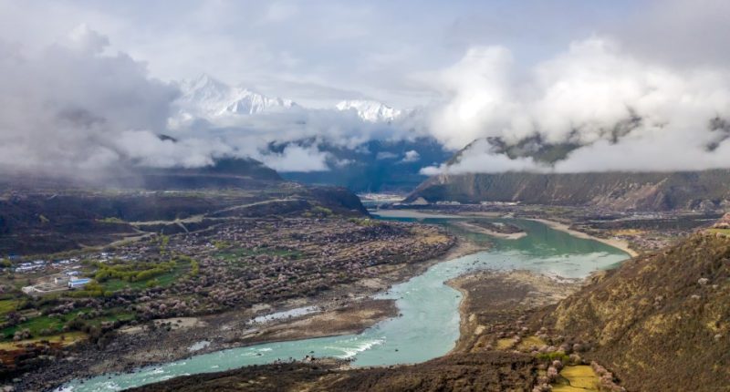 Aerial view of the Yarlung Tsangpo River flowing through a valley with snow-capped mountains in the background.