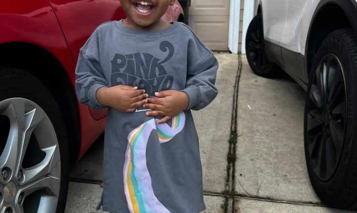 Smiling toddler girl wearing a Pink Floyd shirt and pink shoes.