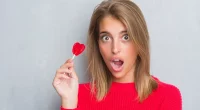 Beautiful young woman over grunge grey wall eating red heart lollipop candy scared in shock with a surprise face, afraid and excited with fear expression