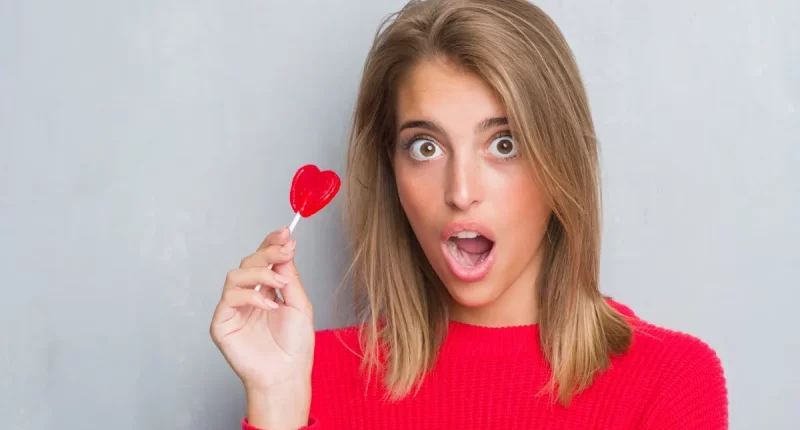 Beautiful young woman over grunge grey wall eating red heart lollipop candy scared in shock with a surprise face, afraid and excited with fear expression