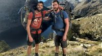 Two men with large backpacks stand on a mountain overlooking a lake.