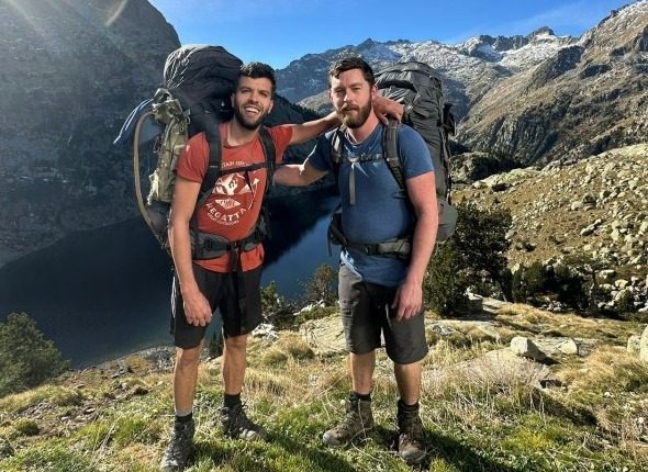 Two men with large backpacks stand on a mountain overlooking a lake.