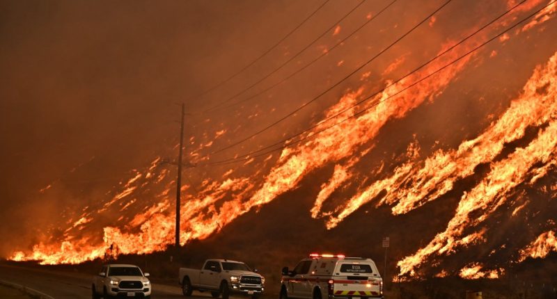Emergency vehicles on a road next to a large wildfire.