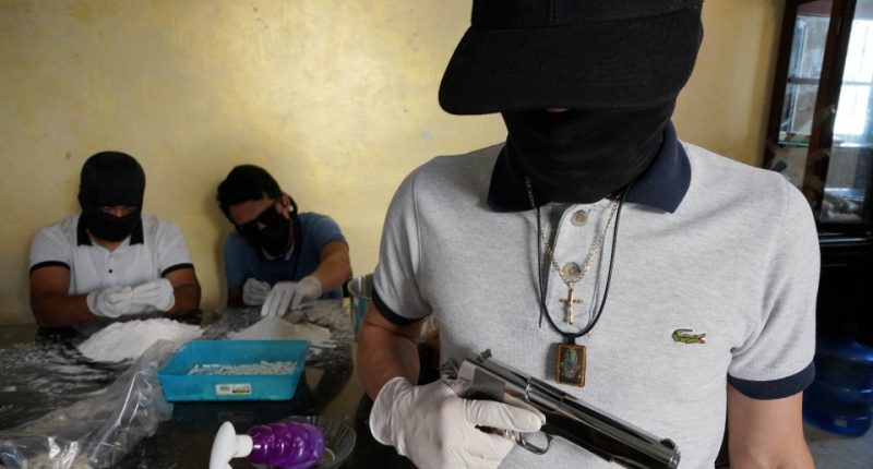 A Sinaloa Cartel member, identified as Guero, holds a handgun while two others prepare drugs.