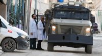 Israeli military vehicle on a street in Jenin, West Bank, with two people in medical scrubs nearby.