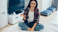 Positive smiling young woman sitting cross-legged on the bed in front of a laptop and holding a cup of coffee