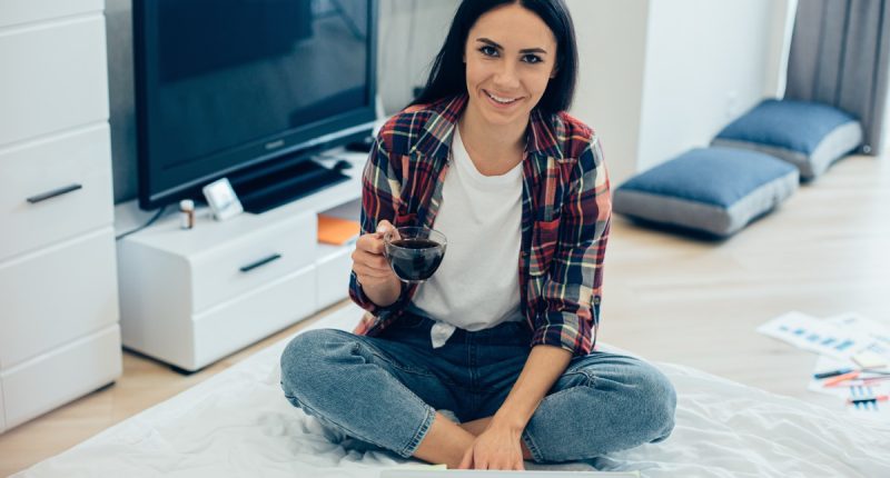 Positive smiling young woman sitting cross-legged on the bed in front of a laptop and holding a cup of coffee