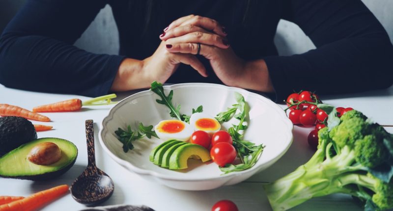 Weight loss plans for women theme white ceramic bowl contained healthy salad (slided avocados, boiled eggs, rocket leaves, tomatoes) on wooden table with blurred woman in black dress / selective focus