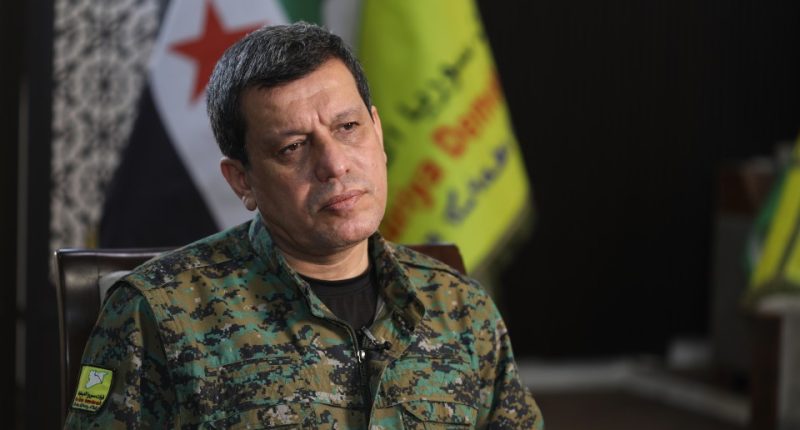 A man in camouflage uniform sits in front of Syrian flags.