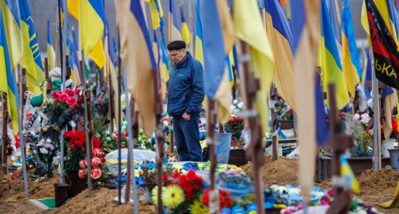 Man mourns at a military cemetery in Kharkiv, Ukraine, among numerous graves marked with Ukrainian flags.
