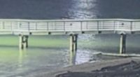 Nighttime view of a pier over water with lights on the far shore and a helicopter in the distance.