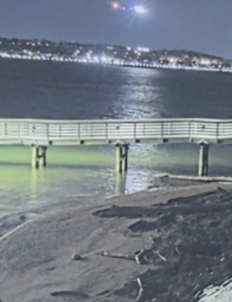 Nighttime view of a pier over water with lights on the far shore and a helicopter in the distance.