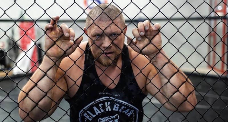 MMA fighter Maxim Novoselov behind a chain-link fence.