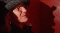 A woman wearing glasses and a hat sits in a dimly lit room.