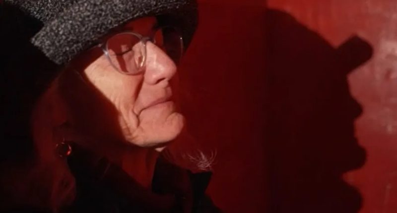 A woman wearing glasses and a hat sits in a dimly lit room.