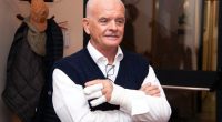 Portrait of a man with his arms crossed, wearing a white shirt and dark blue sweater vest; his right hand is bandaged.