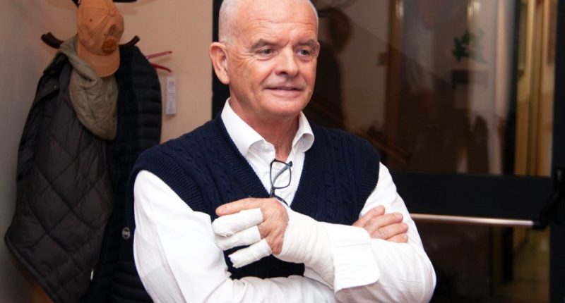 Portrait of a man with his arms crossed, wearing a white shirt and dark blue sweater vest; his right hand is bandaged.
