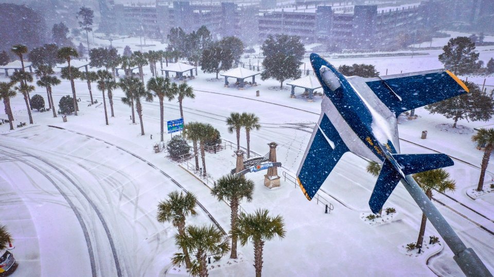Snow falling on palm trees and the Florida Welcome Center, with a Blue Angels plane in the foreground.