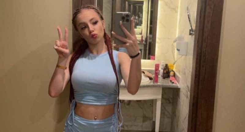 A young woman in a light blue crop top and shorts takes a selfie.