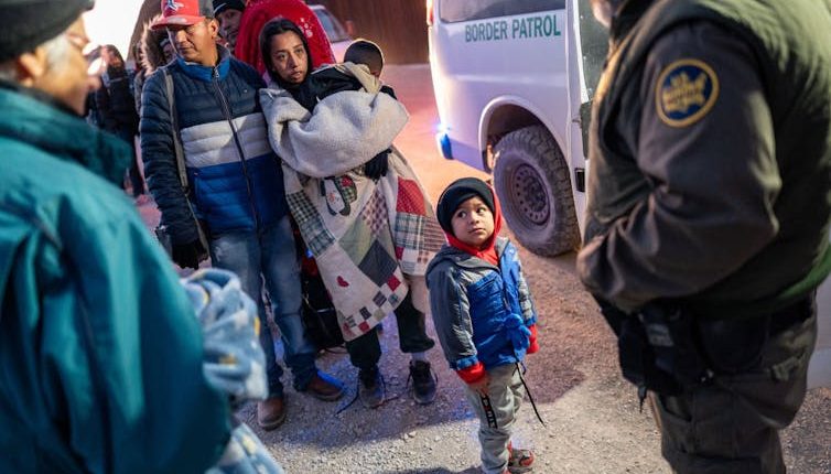 A small boy wearing a jacket stands in front of a woman covered in a blanket and holding a baby. Other people, including a man with a green jacket, stand nearby and next to a white van.