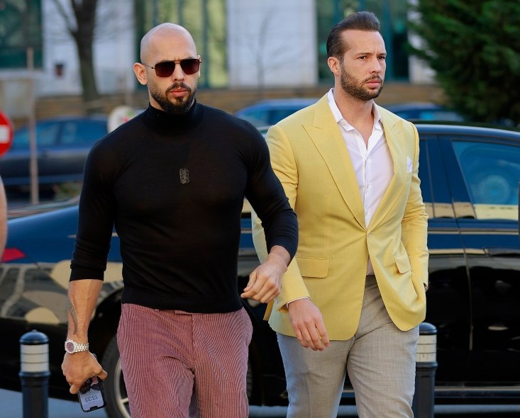 Two men walking outdoors, one in a yellow blazer and the other in a black turtleneck.