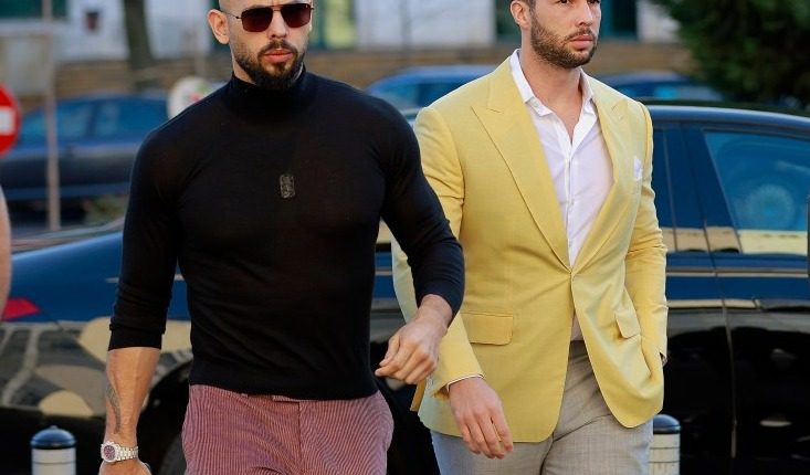 Two men walking outdoors, one in a yellow blazer and the other in a black turtleneck.