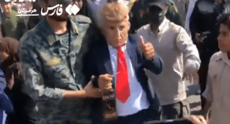 Person in a Donald Trump mask escorted by soldiers.